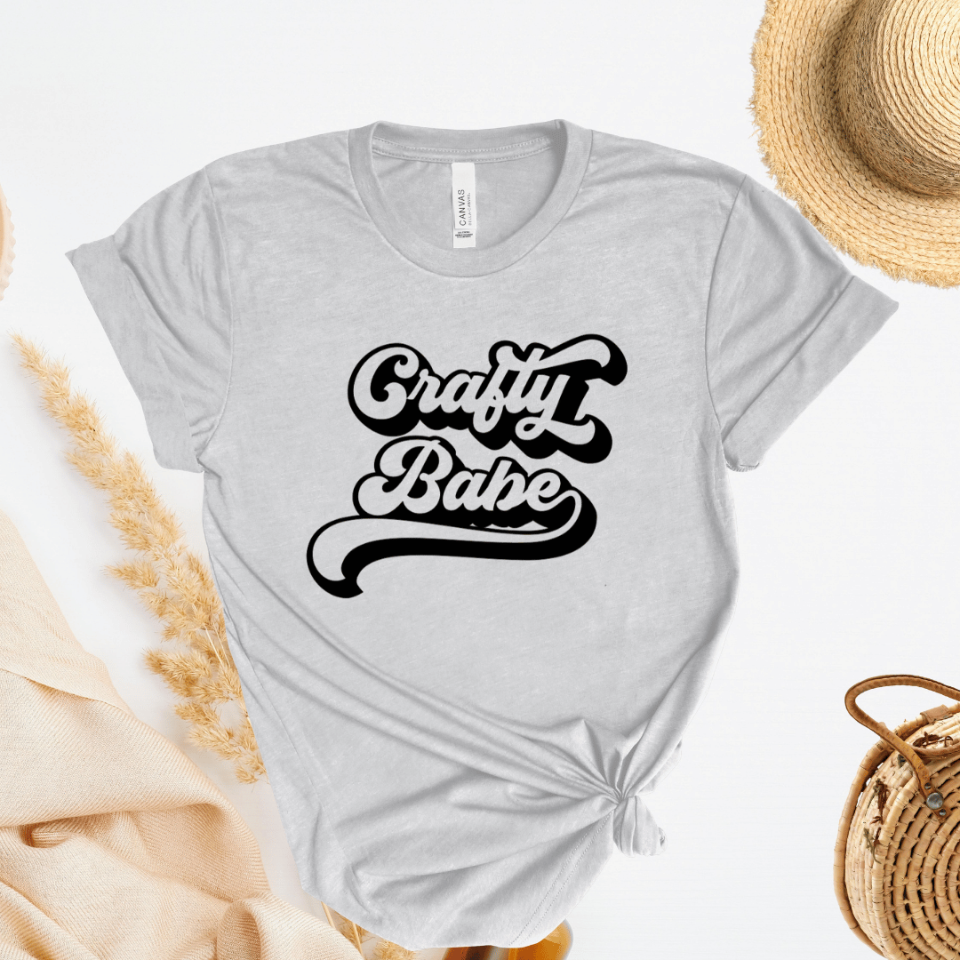 Gray t-shirt with 'Crafty Babe' text, straw hat, and bag on a light background