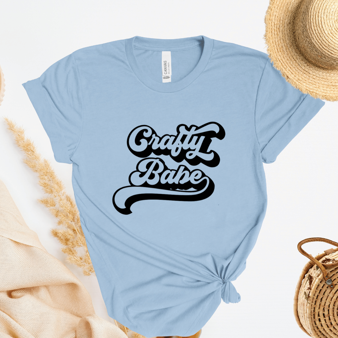 Light blue t-shirt with 'Crafty Babe' text, straw hat, and bag on a light background