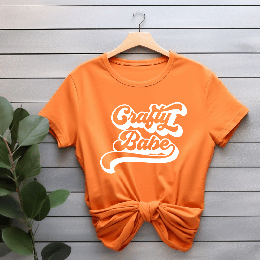Orange t-shirt with 'Crafty Babe' text on a wooden surface with a plant.