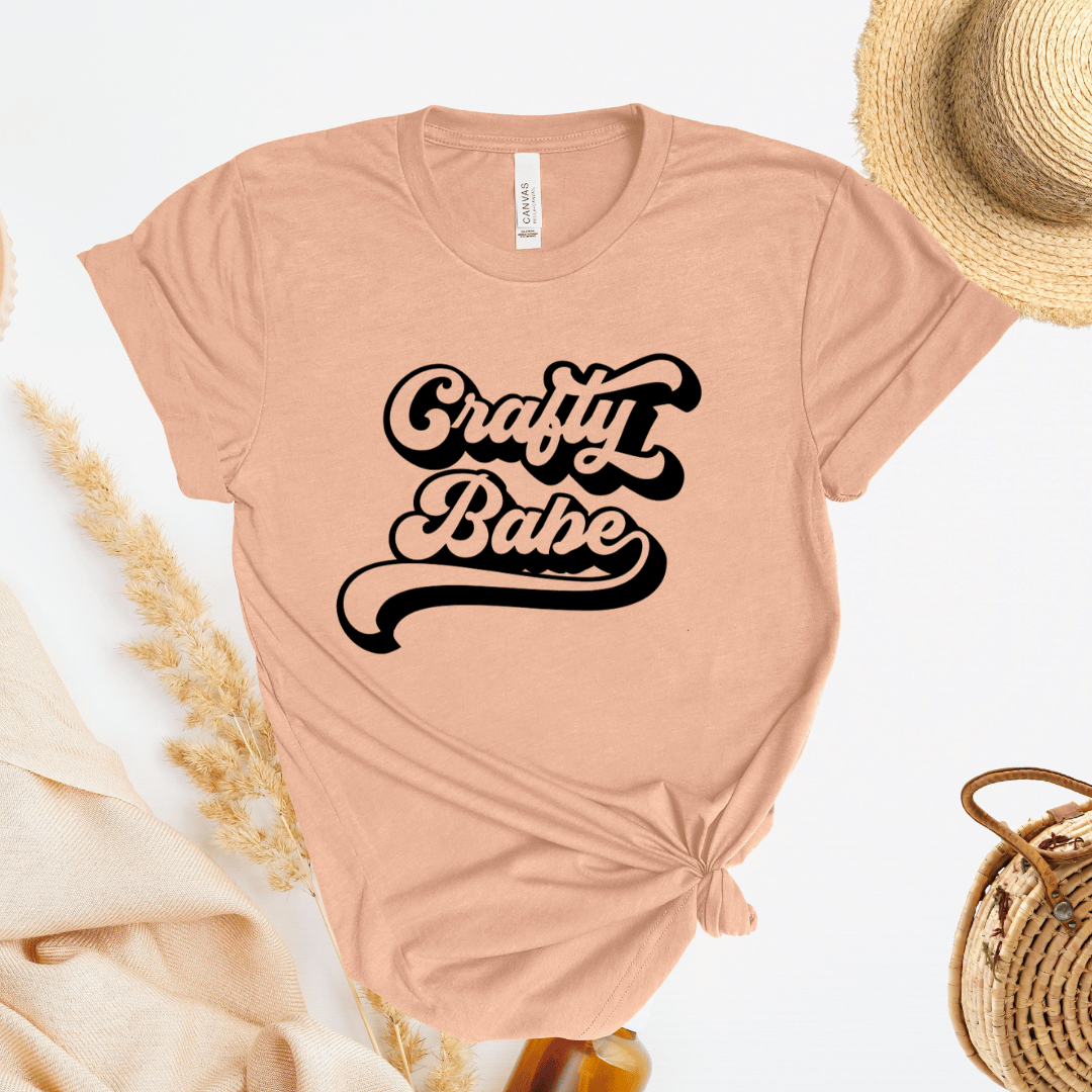 Peach t-shirt with 'Crafty Babe' text, straw hat, and bag on a light background