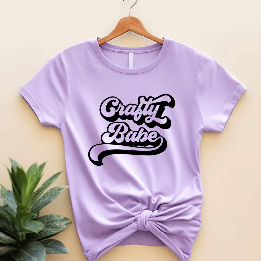 Purple t-shirt with 'Crafty Babe' text on a hanger against a beige background