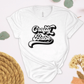White t-shirt with 'Crafty Babe' text on a light background with yarn and flowers.