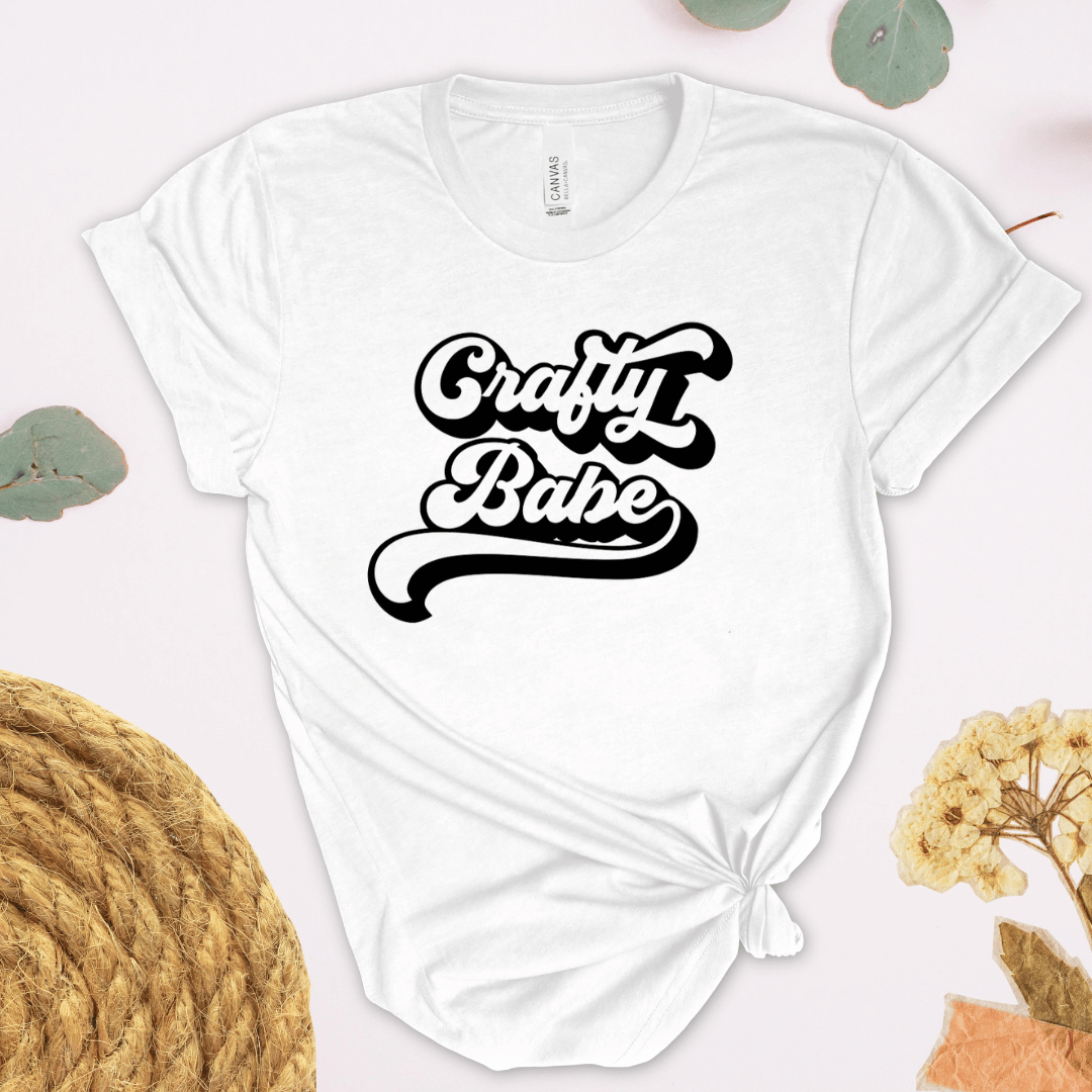 White t-shirt with 'Crafty Babe' text on a light background with yarn and flowers.