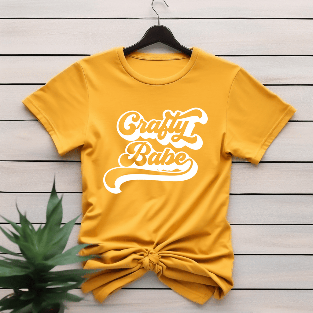 Yellow t-shirt with 'Crafty Babe' text on a wooden surface with a plant.