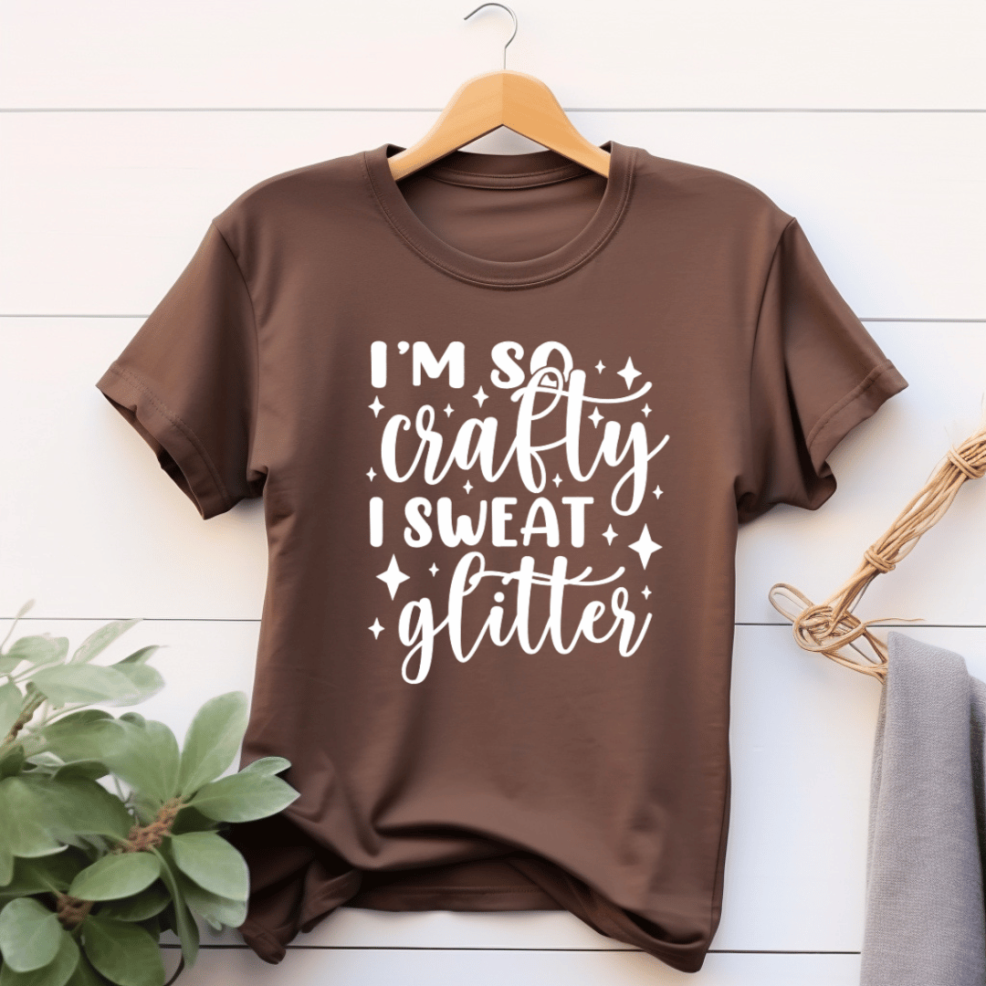 Brown t-shirt with 'I'm so crafty I sweat glitter' text on a wooden hanger against a white wooden background.