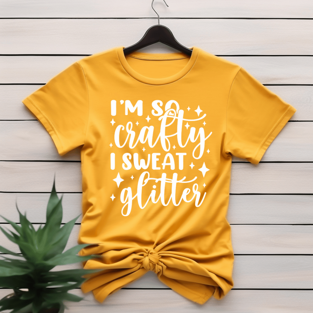 Yellow t-shirt with 'I'm so crafty I sweat glitter' text on a wooden surface.