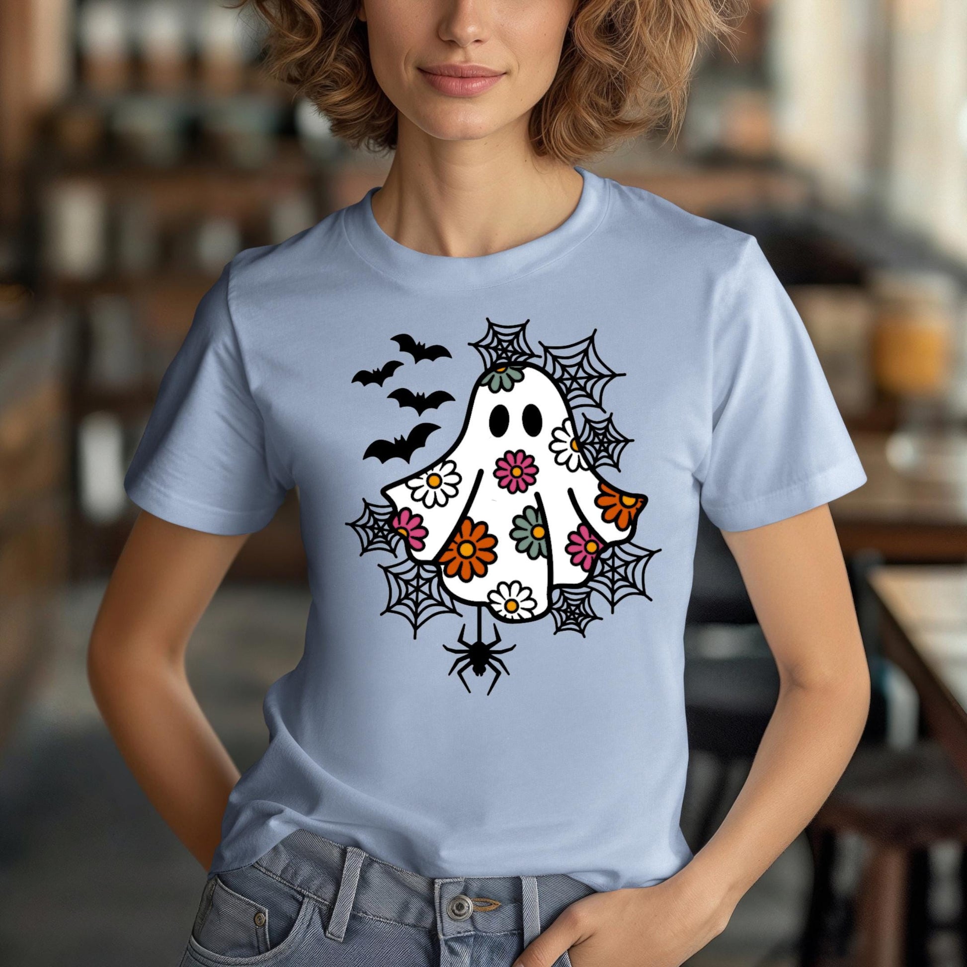 A woman wearing a blue t-shirt with a whimsical design featuring a ghost, bats, and spiders on it.
