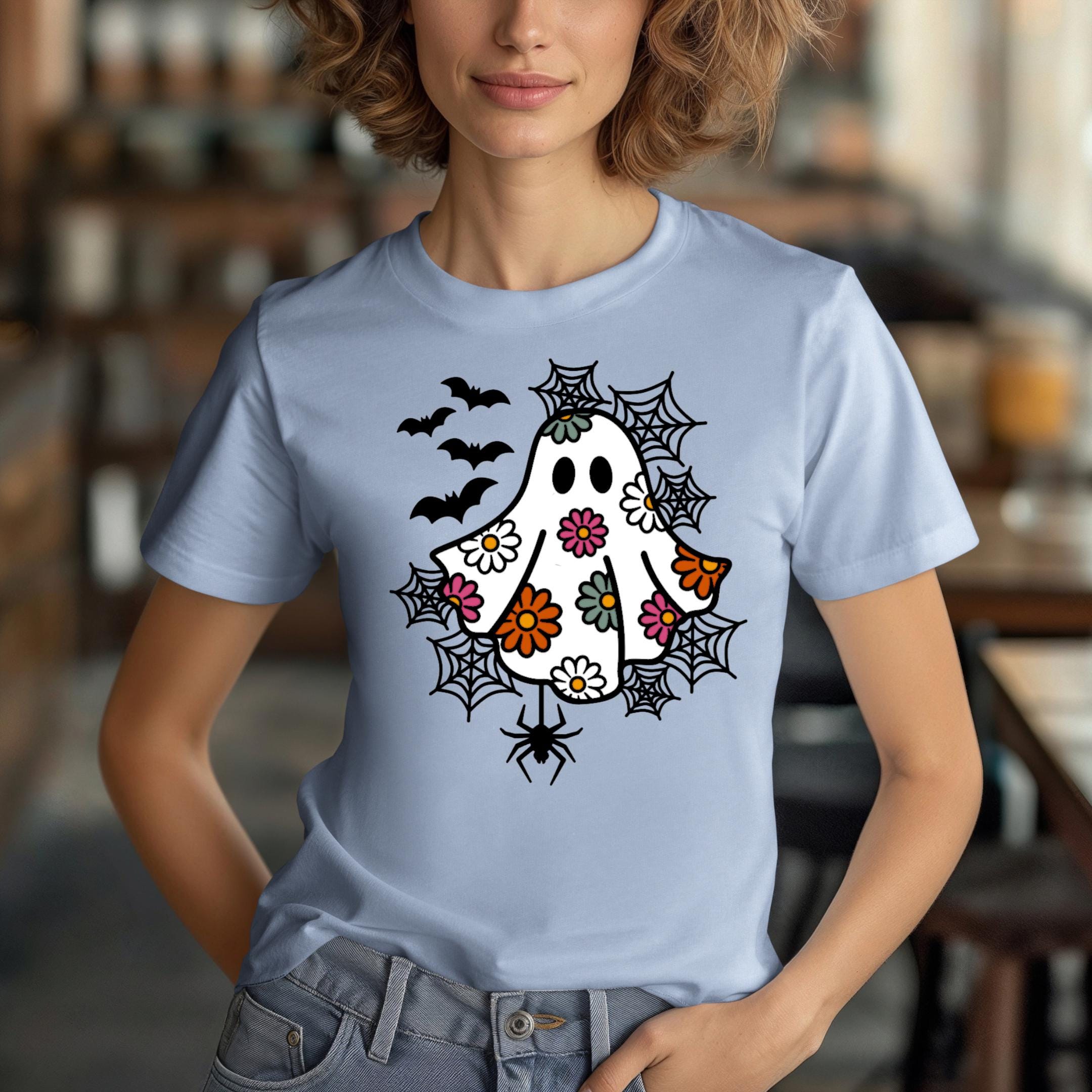 A woman wearing a blue t-shirt with a whimsical design featuring a ghost, bats, and spiders on it.