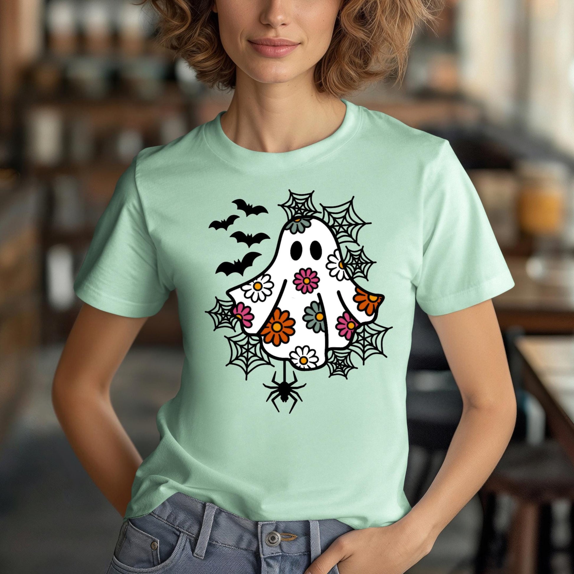 A woman wearing a green t-shirt with a whimsical design featuring a ghost, bats, and spiders on it.
