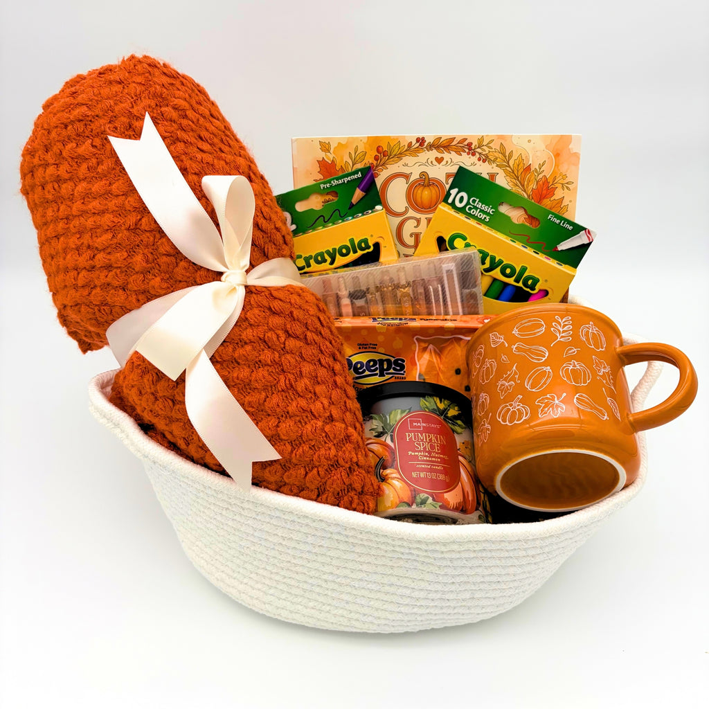 Cream rope fall gift basket with a cozy orange throw blanket, coloring book, markers, pumpkin mug, and candle on white