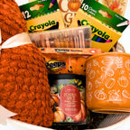 Close-up of the gift basket with a pumpkin spice candle, orange mug with white pumpkin and leaf line art, gel pens, and knit throw
