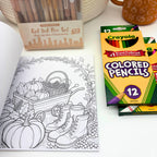 Fall coloring book open to a line drawing of a wooden wagon with pumpkins, pinecones, boots, and a mug framed by a leafy wreath