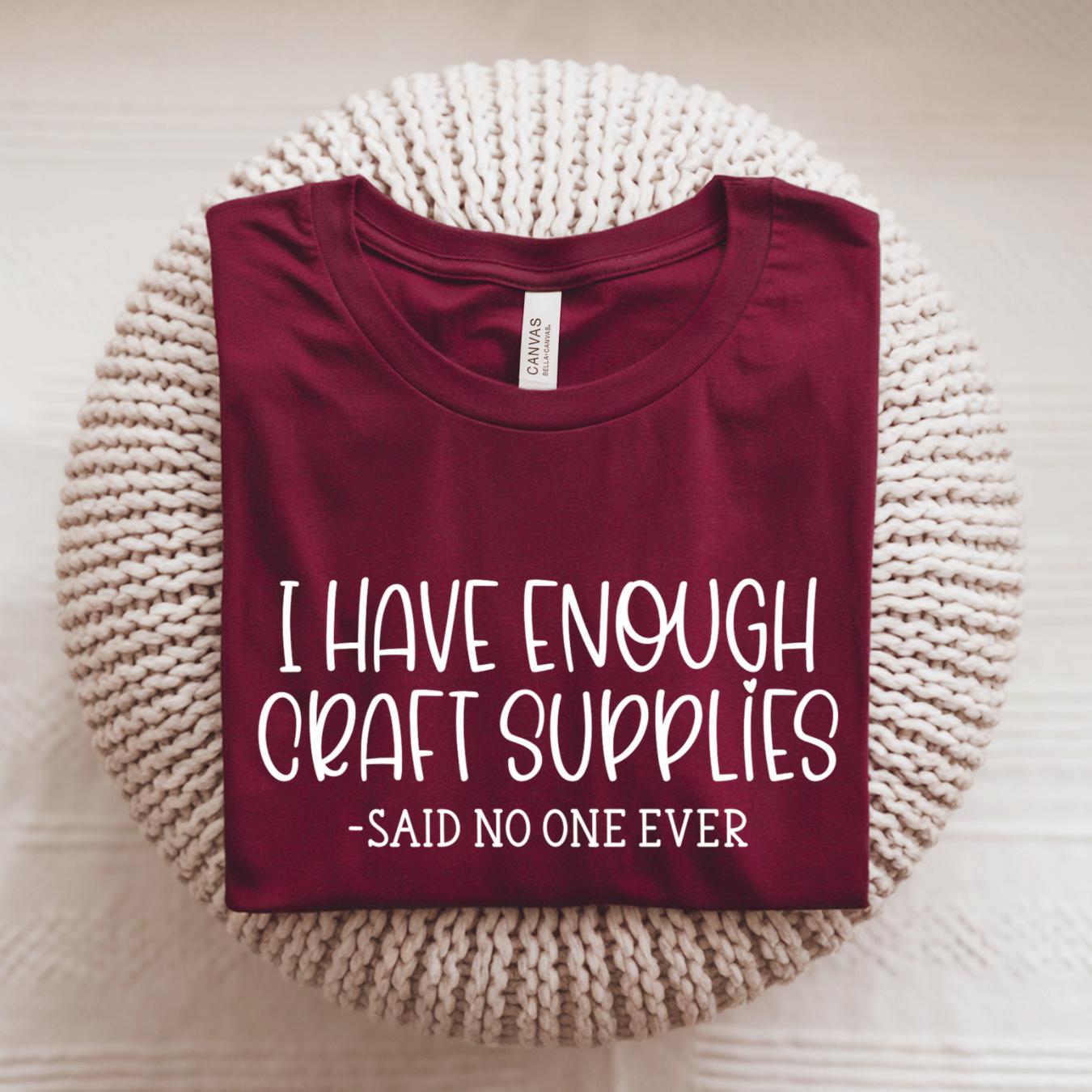 A folded burgundy t-shirt with white text reading 'I HAVE ENOUGH CRAFT SUPPLIES -SAID NO ONE EVER' is displayed on a round, cream-colored knitted pouf. 