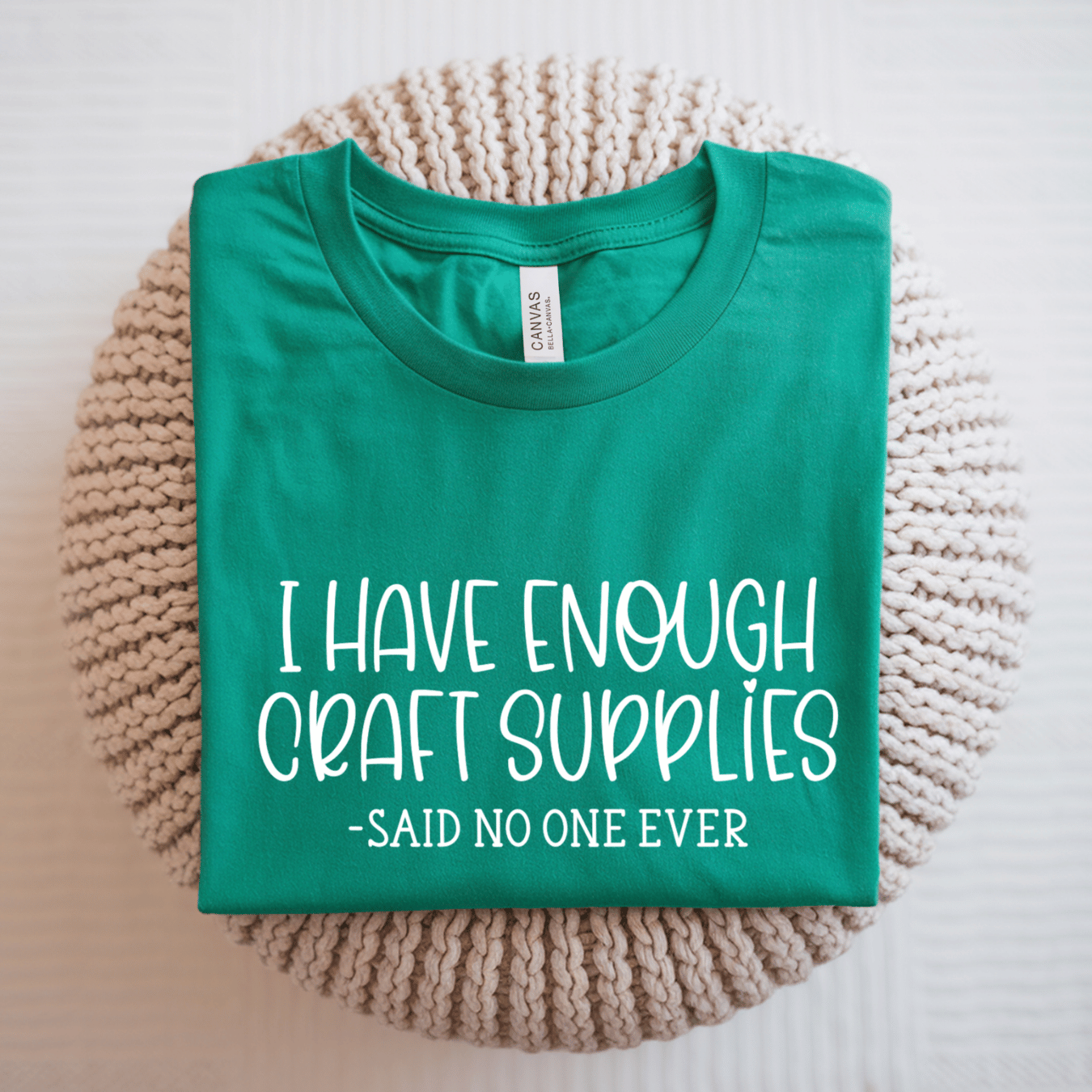 A folded kelly green t-shirt with white text reading 'I HAVE ENOUGH CRAFT SUPPLIES -SAID NO ONE EVER' is displayed on a round beige knit placemat against a white background. 