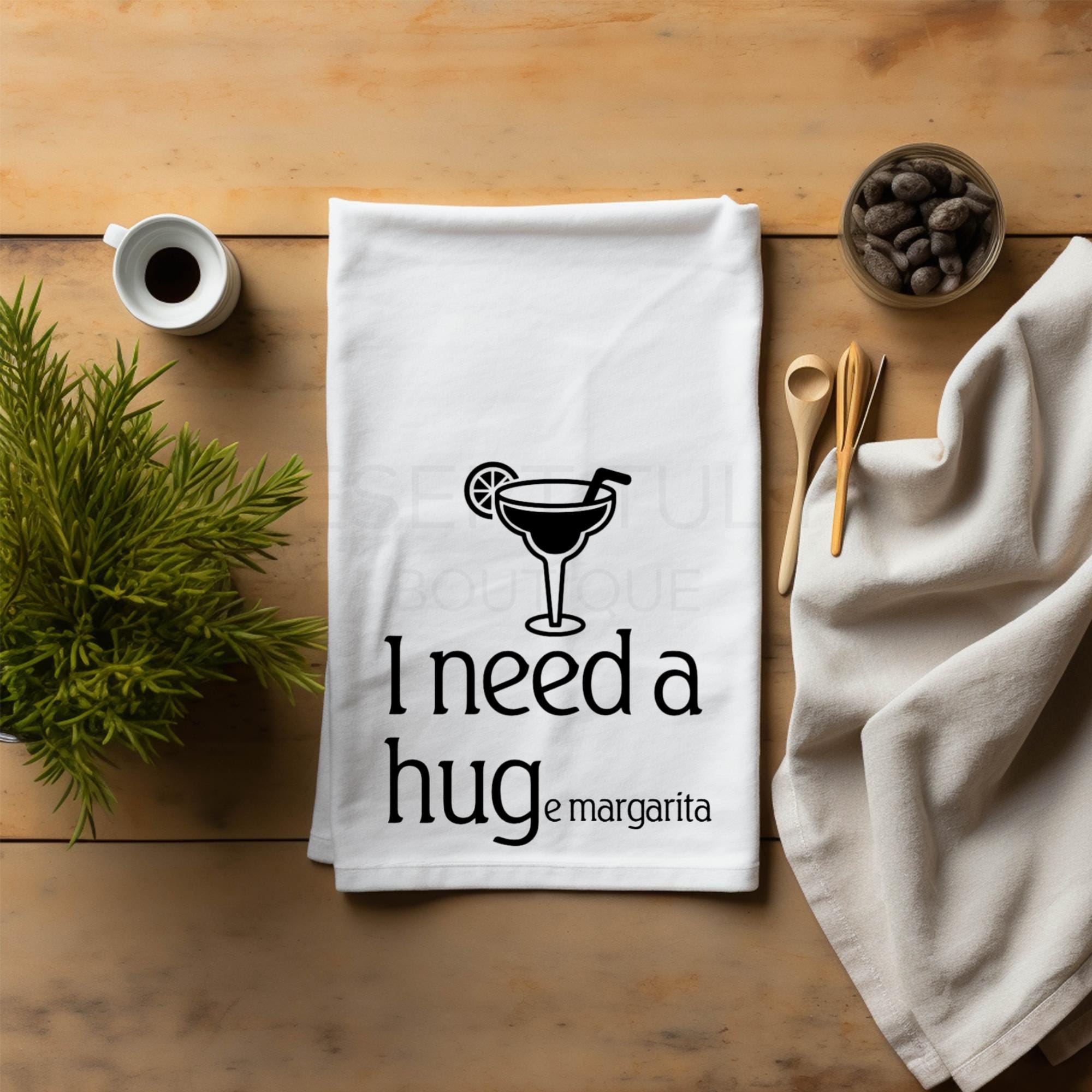 A white cotton flour sack kitchen tea towel with black text and graphic design lying on a wooden table, featuring the phrase 'I need a hug(e margarita)' displayed in a casual kitchen setting with herbs and kitchen utensils.