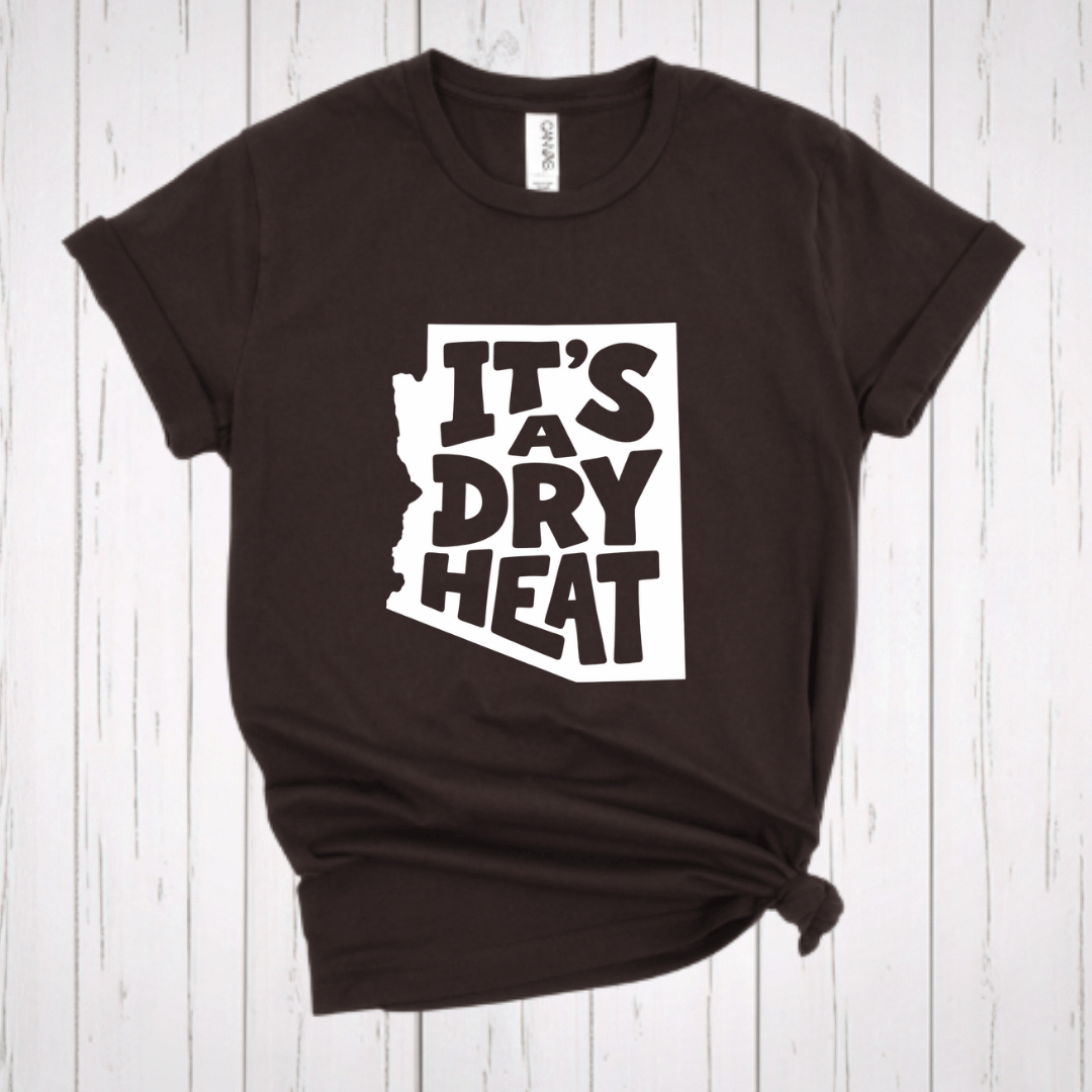 A dark brown t-shirt with a white graphic containing the text 'IT'S A DRY HEAT' in the shape of Arizona, laid flat on a rustic wooden background.