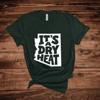 A dark green t-shirt with a white graphic containing the text 'IT'S A DRY HEAT' in the shape of Arizona, laid flat on a rustic wooden background.