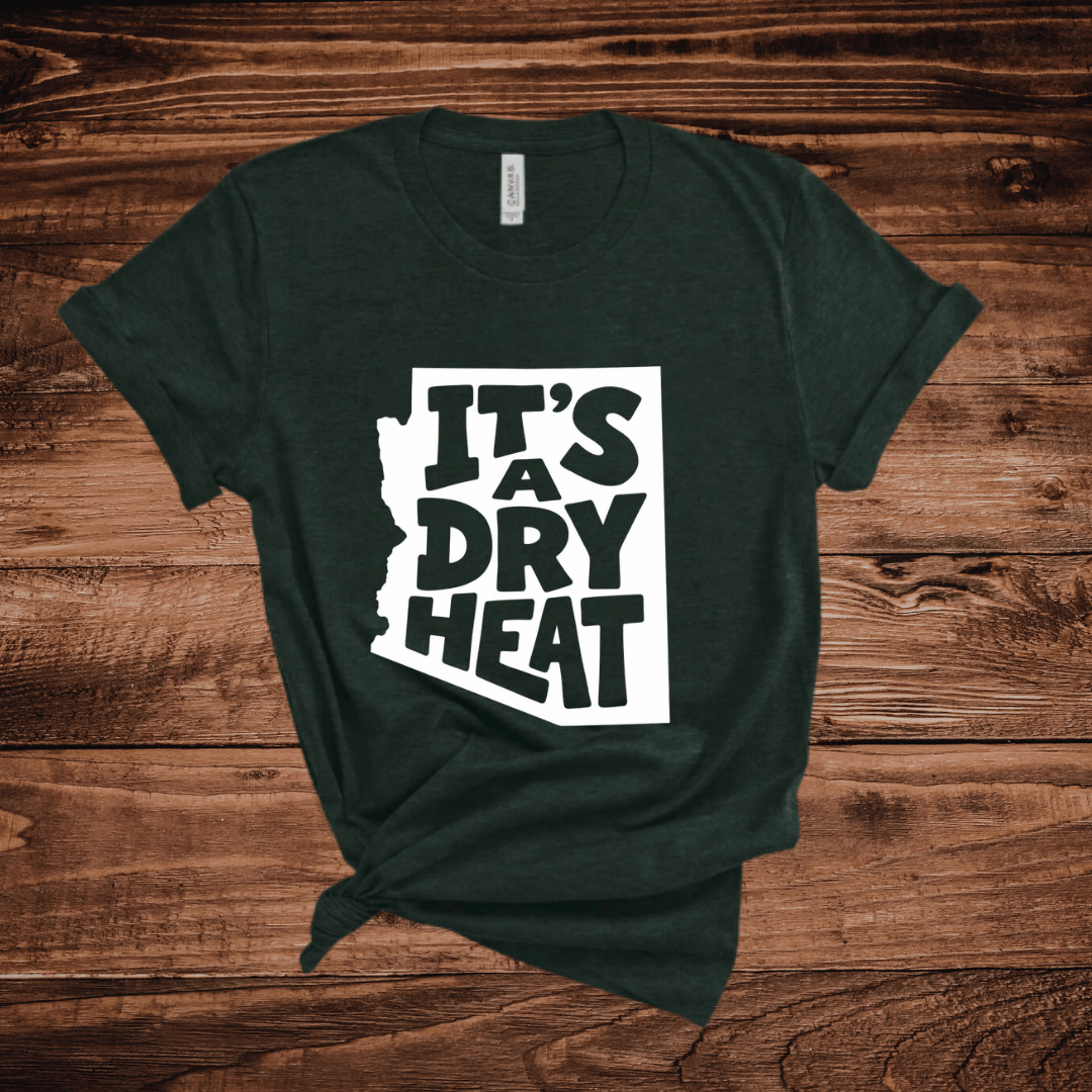 A dark green t-shirt with a white graphic containing the text 'IT'S A DRY HEAT' in the shape of Arizona, laid flat on a rustic wooden background.