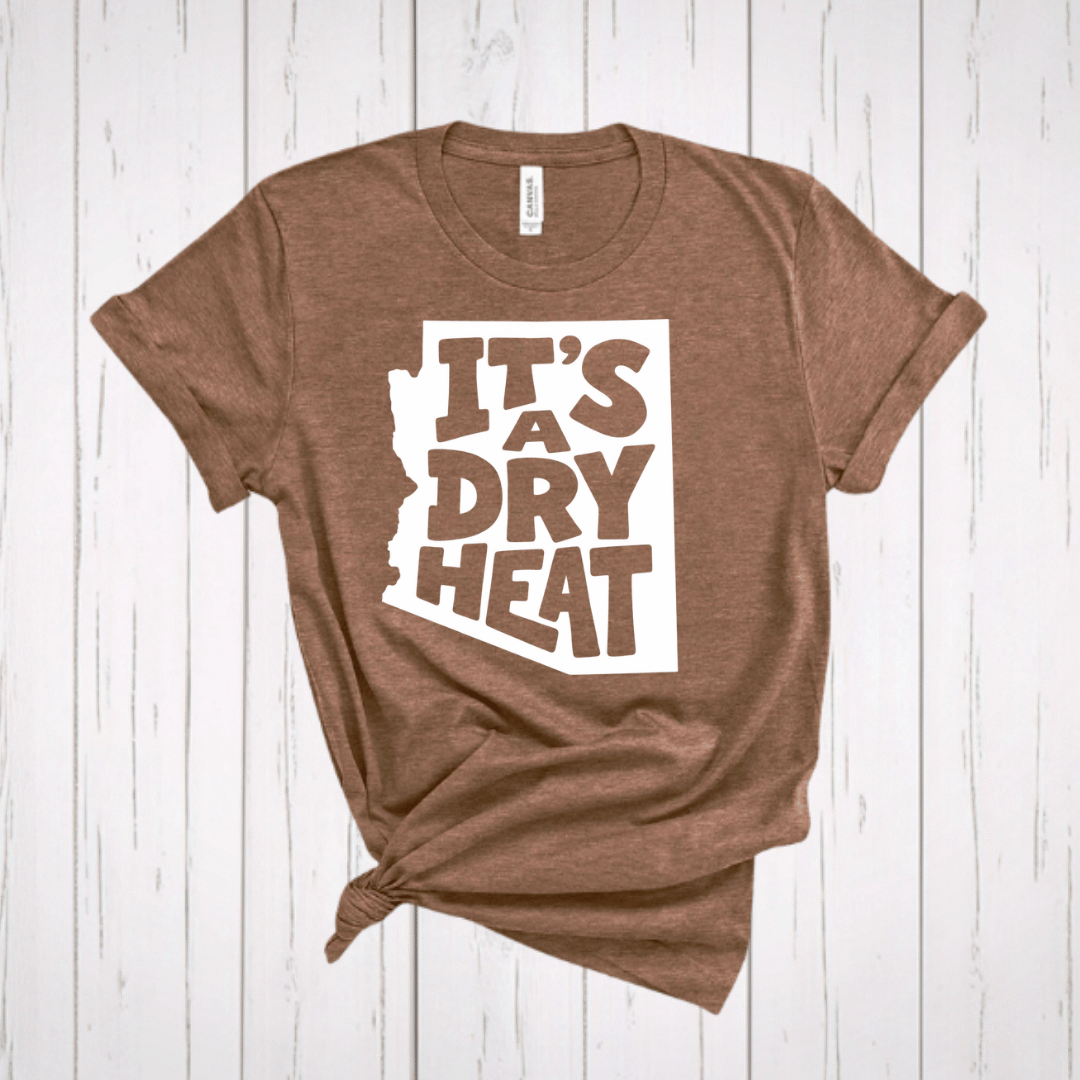 A light brown t-shirt with a white graphic containing the text 'IT'S A DRY HEAT' in the shape of Arizona, laid flat on a rustic wooden background.