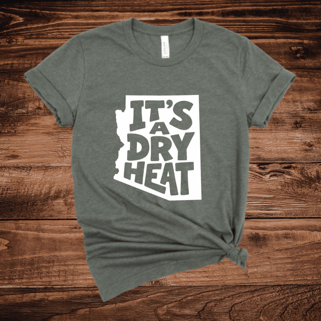 A light green t-shirt with a white graphic containing the text 'IT'S A DRY HEAT' in the shape of Arizona, laid flat on a rustic wooden background.