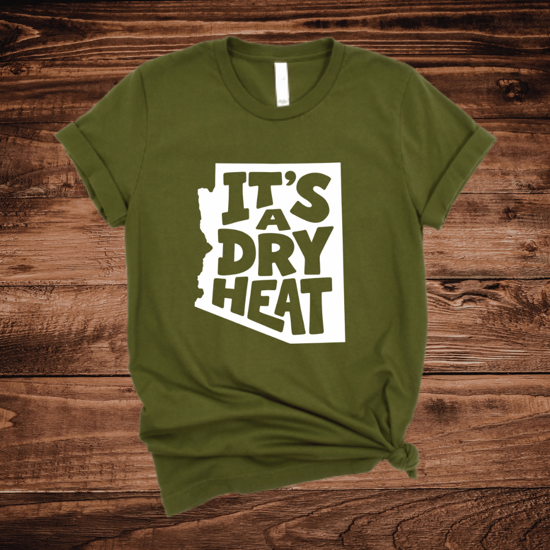 An olive green t-shirt with a white graphic containing the text 'IT'S A DRY HEAT' in the shape of Arizona, laid flat on a rustic wooden background.