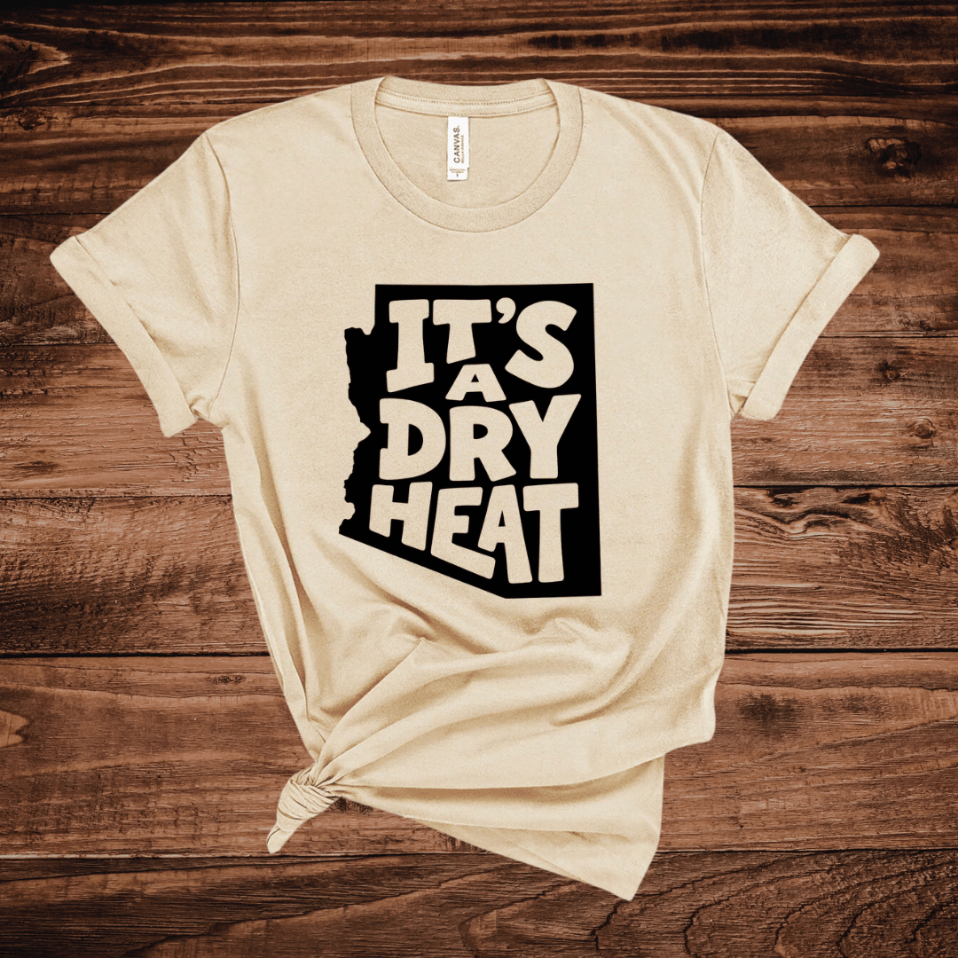 A tan t-shirt with a white graphic containing the text 'IT'S A DRY HEAT' in the shape of Arizona, laid flat on a rustic wooden background.