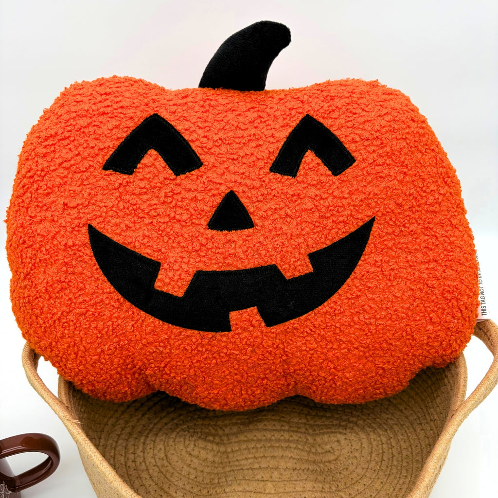Close-up of an orange boucle jack-o'-lantern pillow with a black appliqué face and stem sitting on top of a tan rope basket