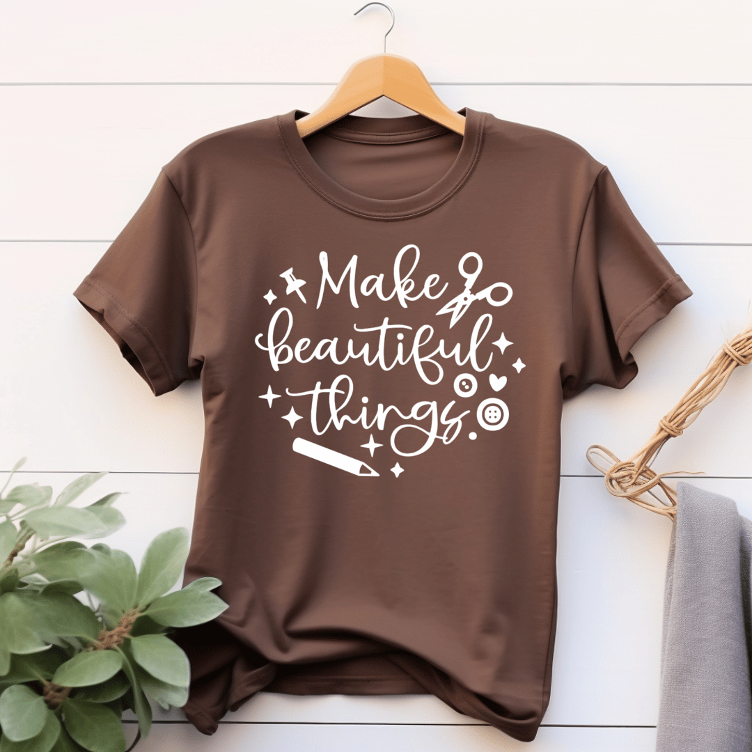 Brown t-shirt with 'Make beautiful things' text on a wooden hanger against a white wooden background.