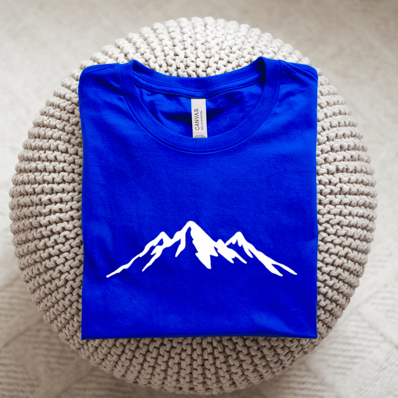 A folded royal blue t-shirt with a simple white mountain graphic on the front, displayed on a round, textured beige knit pouf. 
