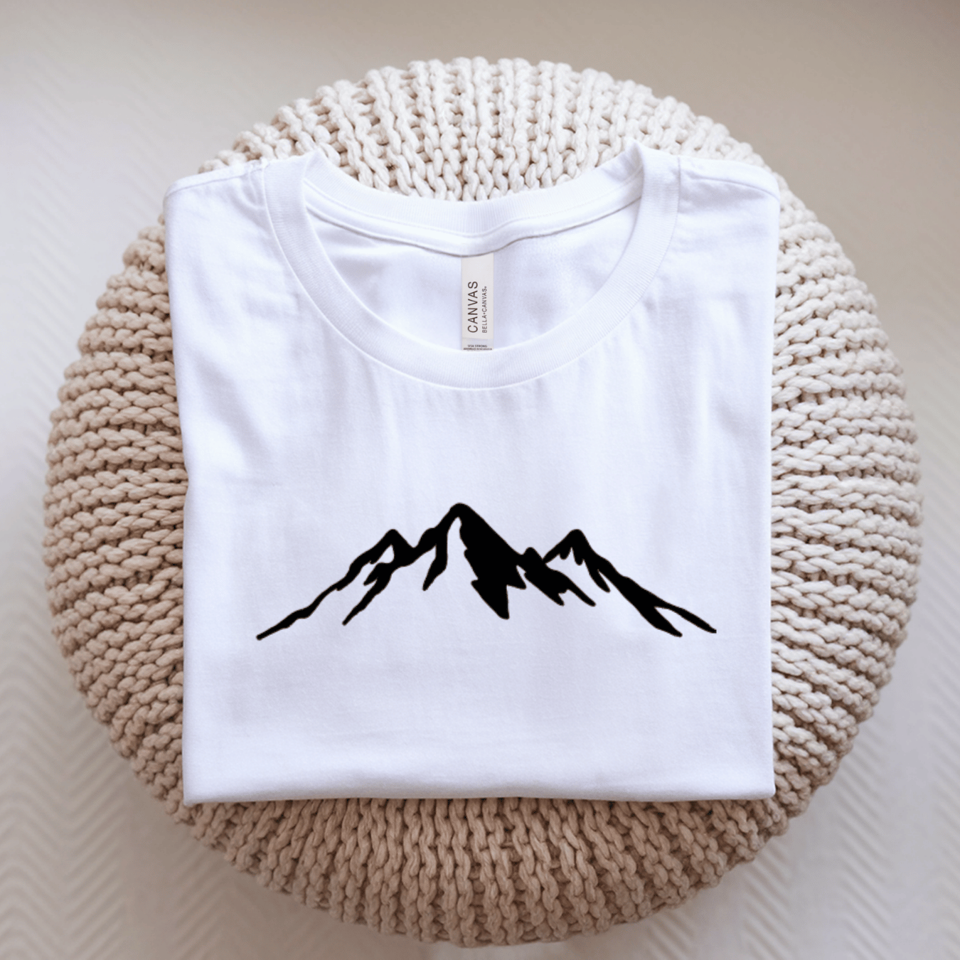 A folded white t-shirt with a black mountain silhouette graphic on the chest, displayed on a round beige knitted cushion.