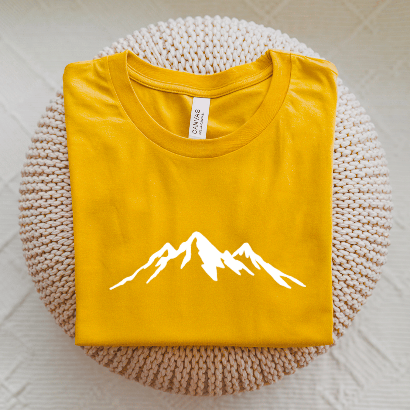 A folded yellow t-shirt with a white mountain graphic on the front, displayed on a round beige knit cushion over a light textured background.