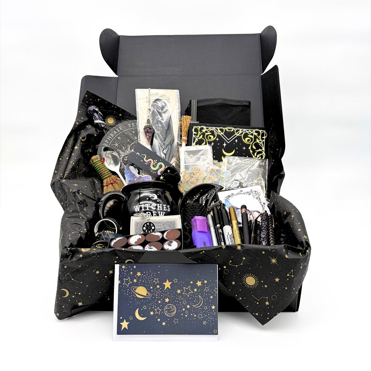 Open black gift box with celestial tissue holding the mystical witchy gift box contents, with a gold star and moon greeting card in front