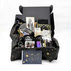 Open black gift box with celestial tissue holding the mystical witchy gift box contents, with a gold star and moon greeting card in front