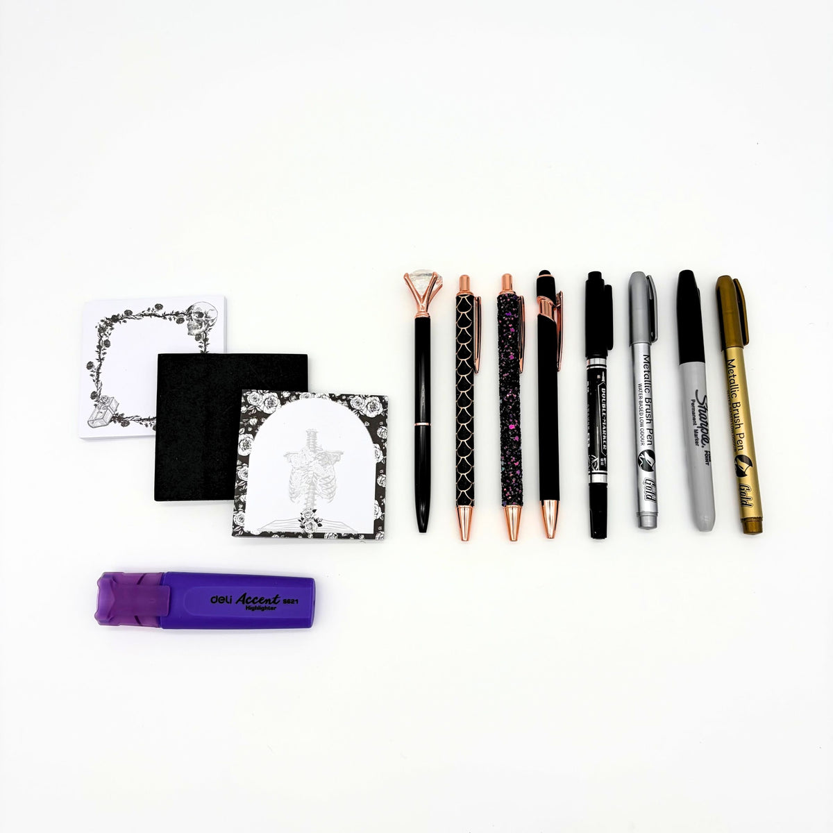 Eight pens and markers from the mystical witchy gift box beside skull and ribcage sticky note pads and a purple highlighter