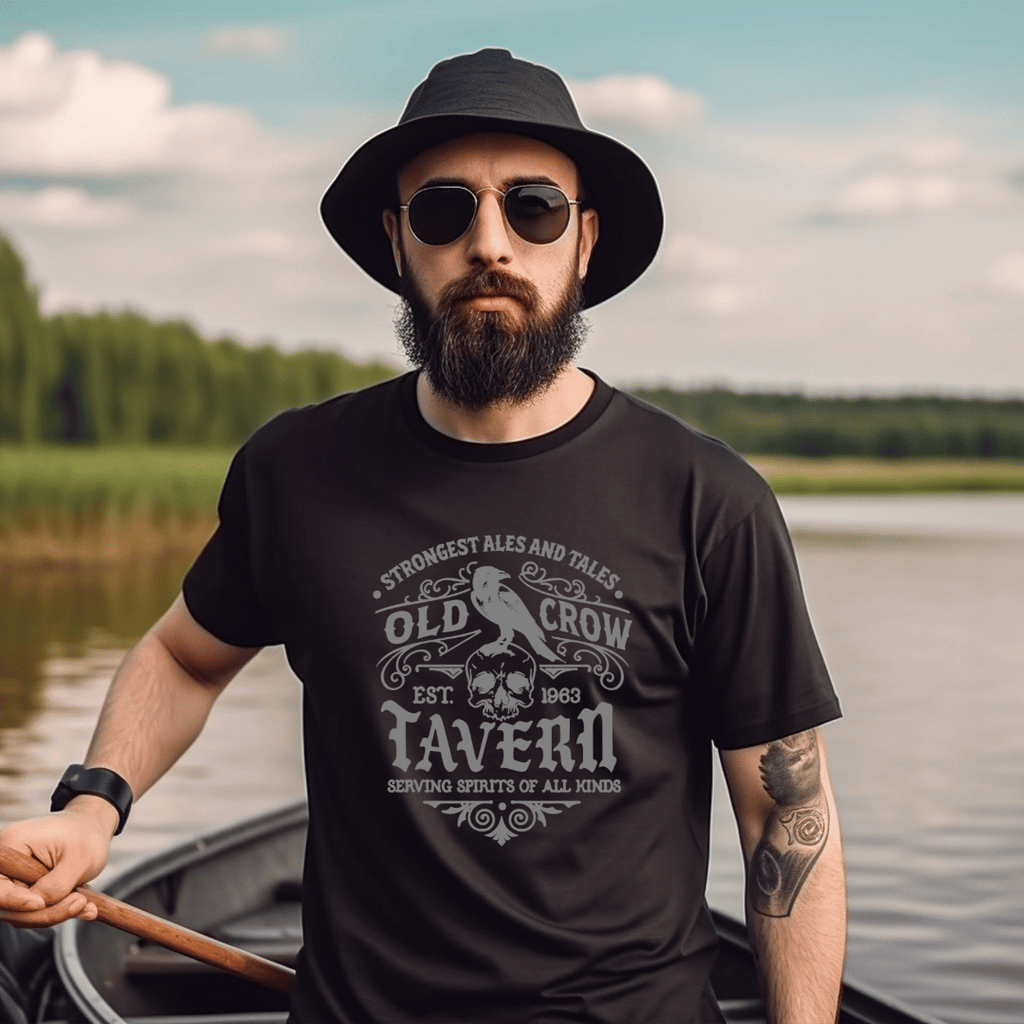 Man wearing an 'Old Crow Tavern' black t-shirt with a vintage tavern design, standing by a lake.