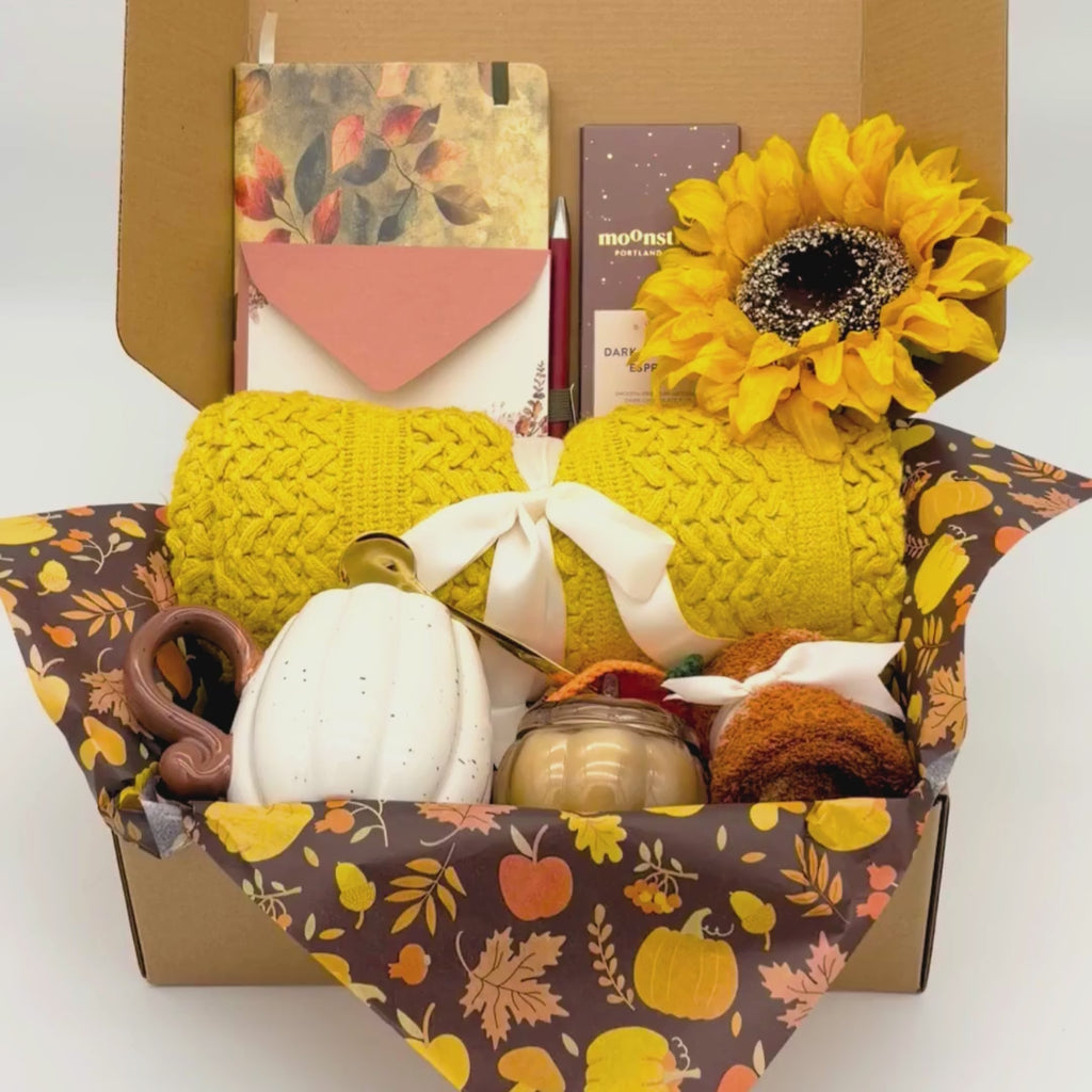 Overhead video of a cozy fall gift box to reveal the knit throw blanket, pumpkin mug, candle and fall treats inside