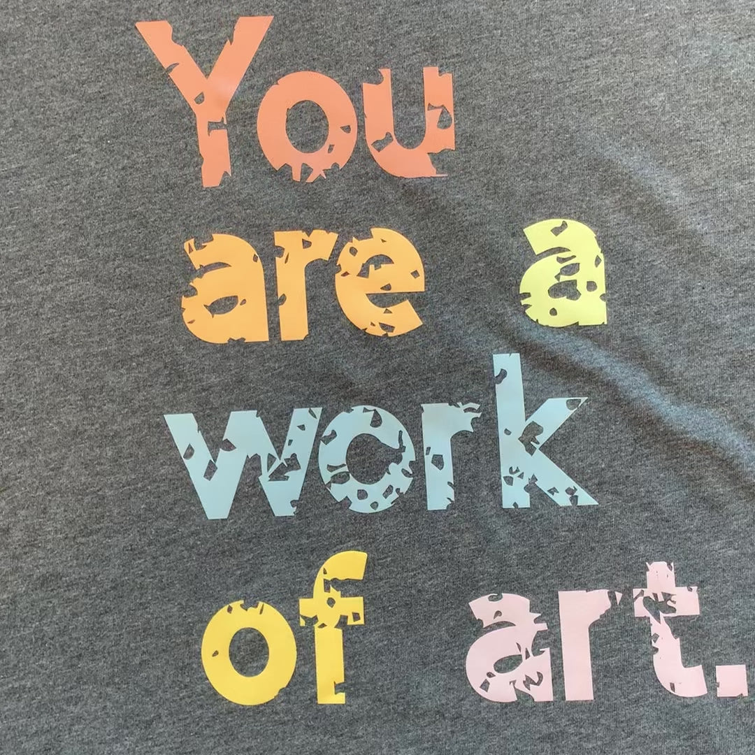 Video of a grey t-shirt with the text 'You are a work of art' printed in colorful letters.