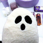 Close-up of a fluffy white sherpa ghost pillow with black oval eyes and an open mouth, with the tumbler and candle behind it