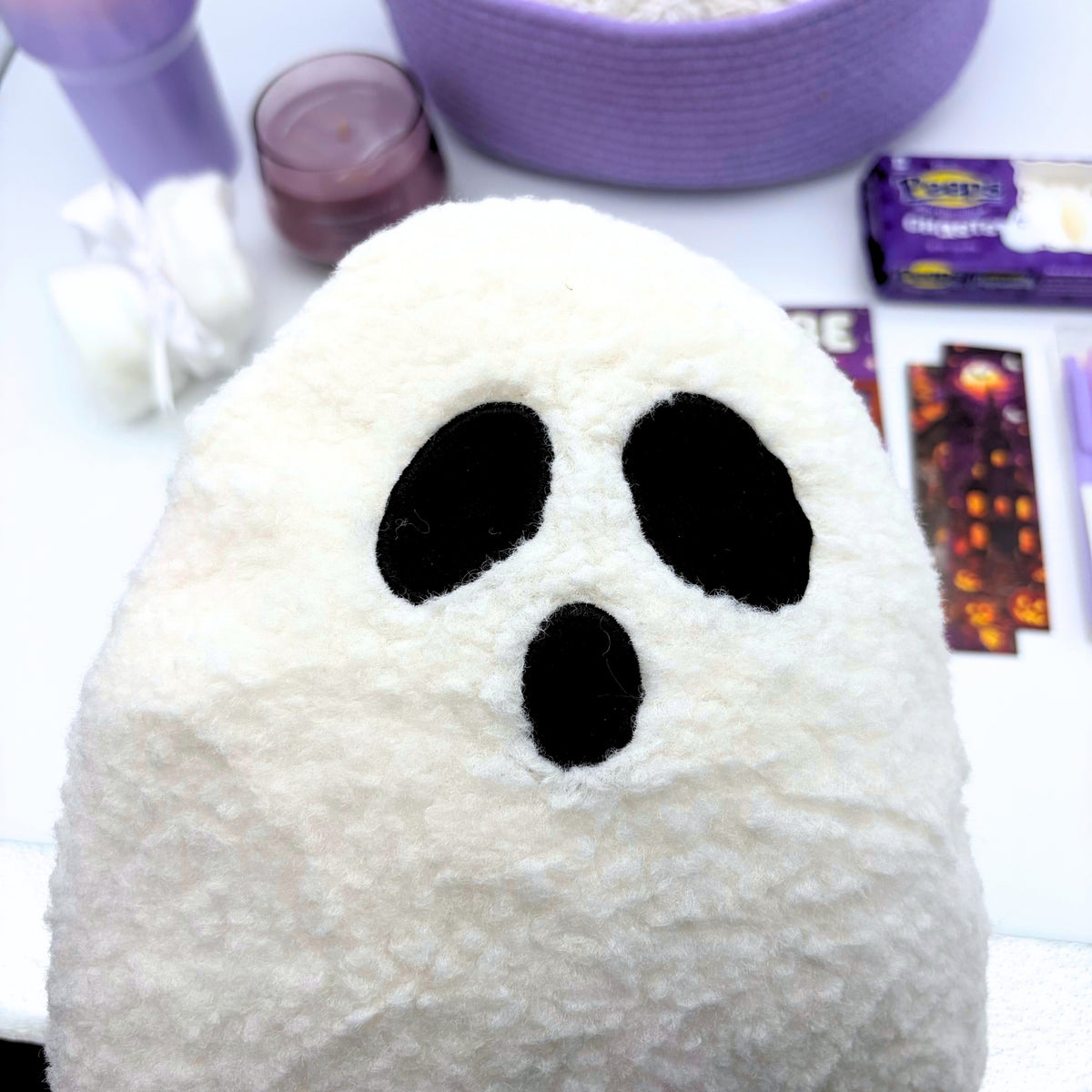 Close-up of a fluffy white sherpa ghost pillow with black oval eyes and an open mouth, with the tumbler and candle behind it