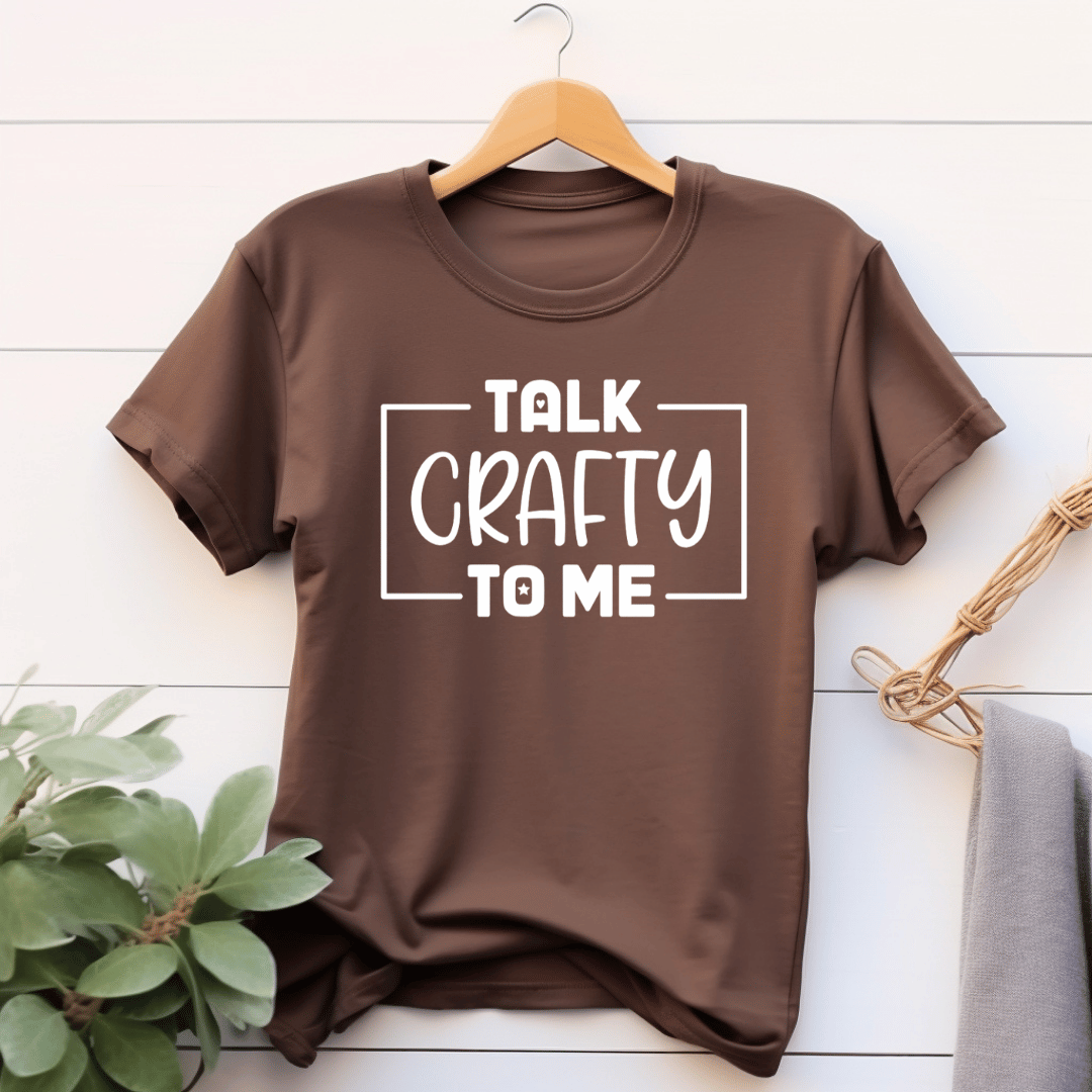 Brown t-shirt with 'Talk Crafty to Me' text on a wooden hanger against a white wooden background.