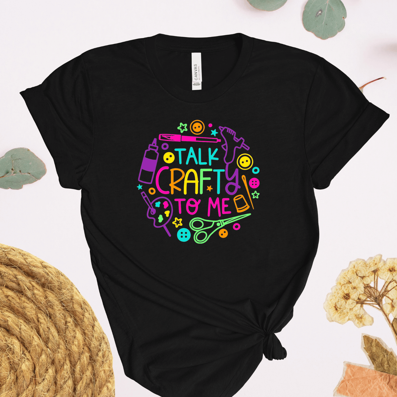 A black short-sleeve t-shirt with a colorful graphic that reads 'TALK CRAFTY TO ME' surrounded by craft-themed icons like scissors, paintbrushes, and beads. The shirt is styled in a flat-lay with decorative elements.