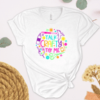 A white short-sleeve t-shirt with a colorful graphic that reads 'TALK CRAFTY TO ME' surrounded by craft-themed illustrations, styled in a flat-lay design.