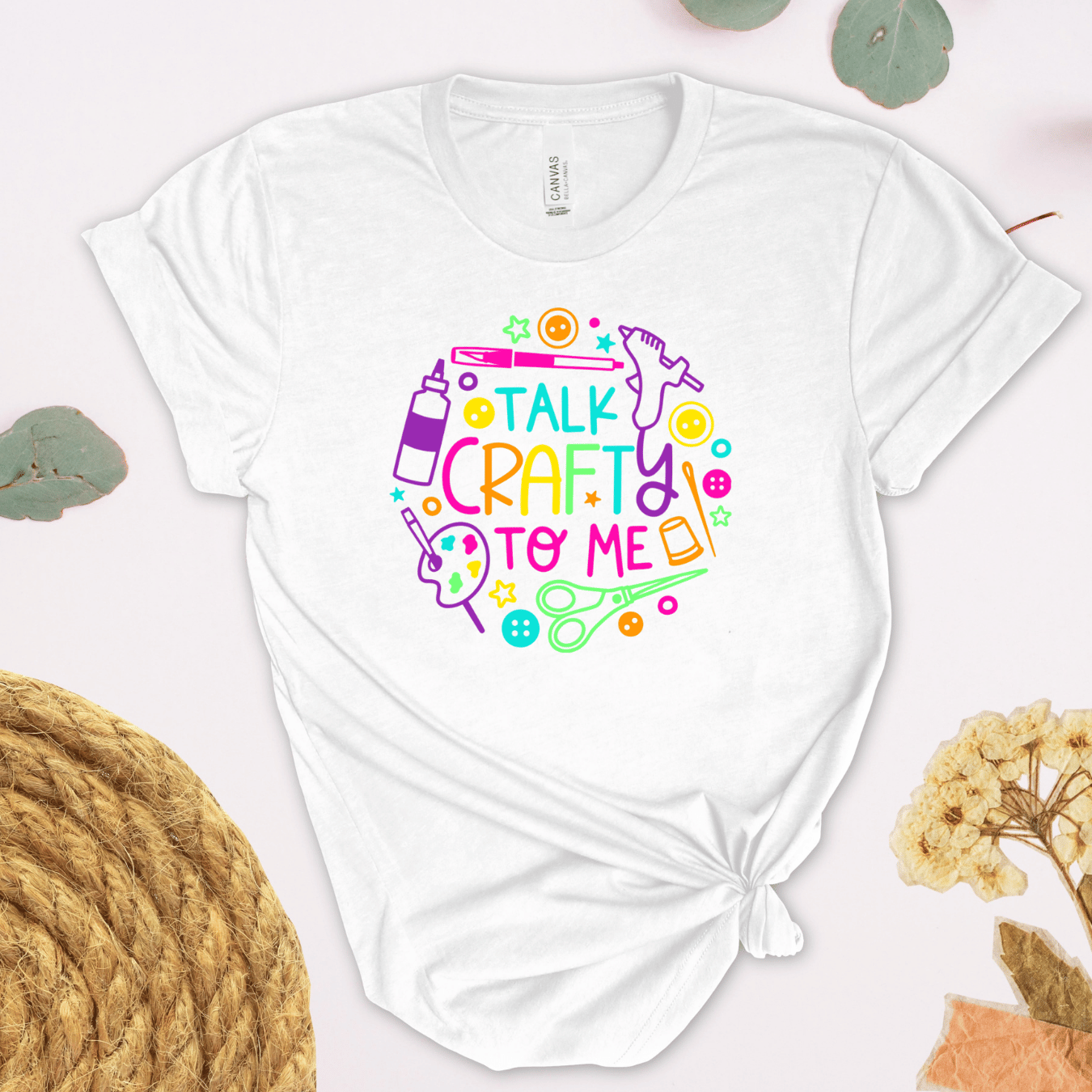 A white short-sleeve t-shirt with a colorful graphic that reads 'TALK CRAFTY TO ME' surrounded by craft-themed illustrations, styled in a flat-lay design.