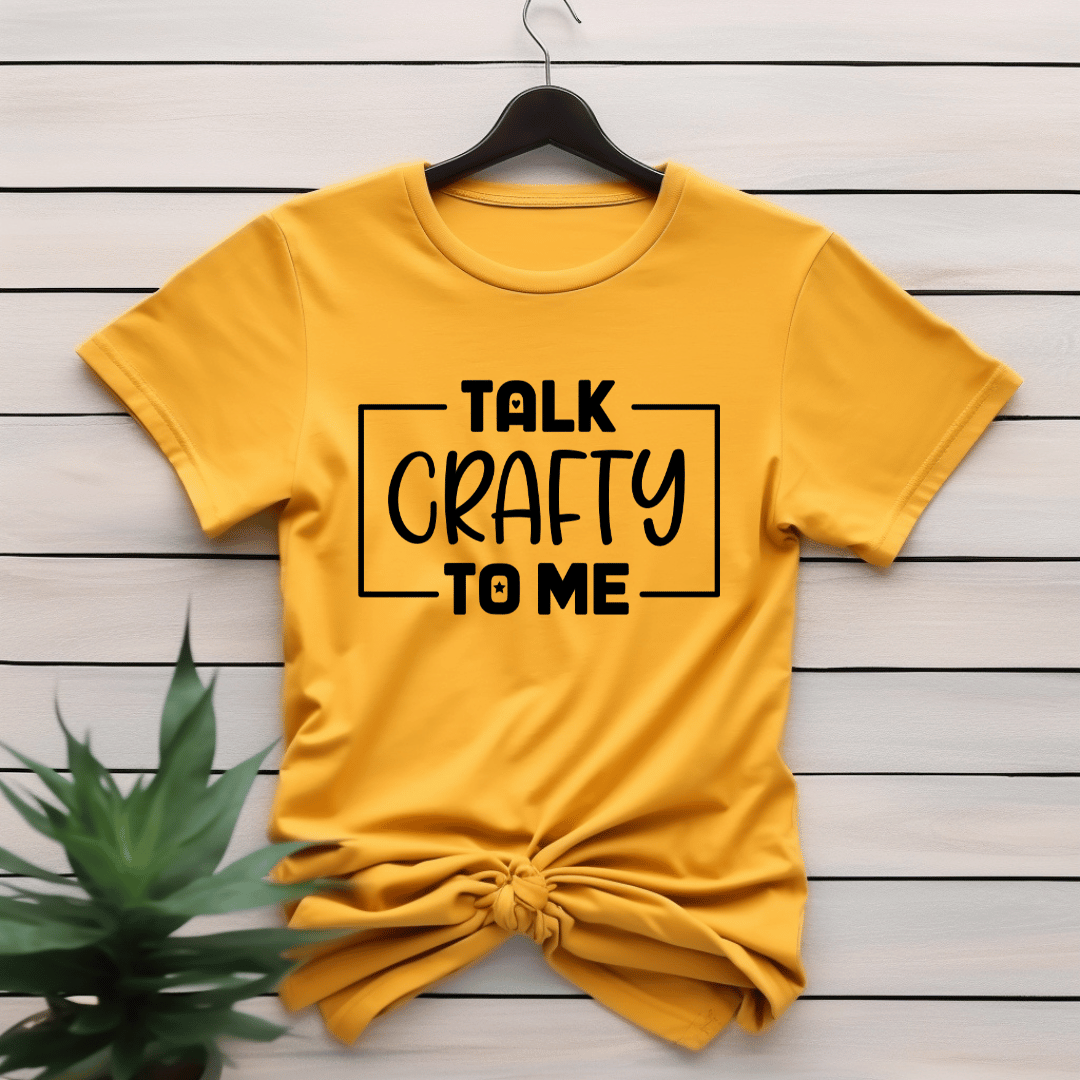 Yellow t-shirt with 'Talk Crafty to Me' text on a wooden surface with a plant.