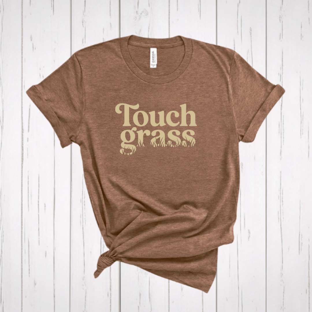 Touch Grass funny meme t-shirt with retro serif font and grass blade lettering on a light brown shirt